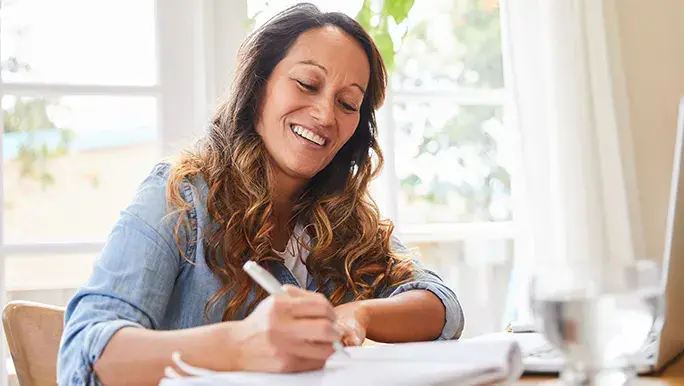 A woman is sitting down at the table and smiling while she journals in her self-care diary.