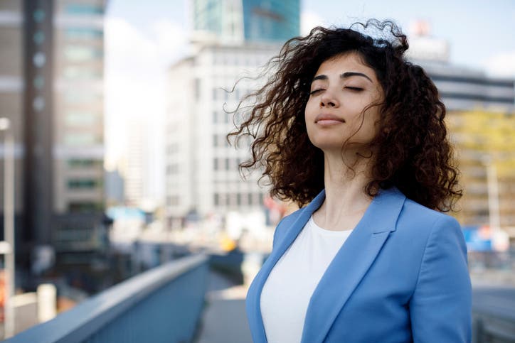 Dark-haired woman with a blue jacket is standing in a city with her eyes closed, practising mindfulness