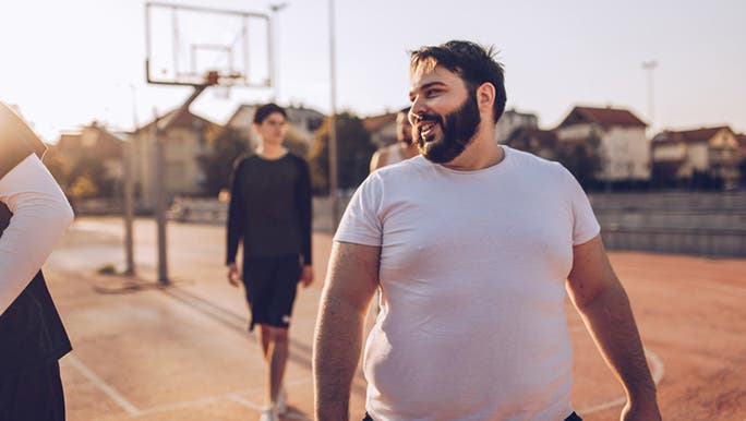 A man with dark hair and a beard wearing a white T-shirt is walking on a basketball court with friends and smiling
