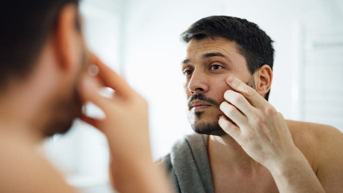 A young man is looking at his skin in a bathroom mirror