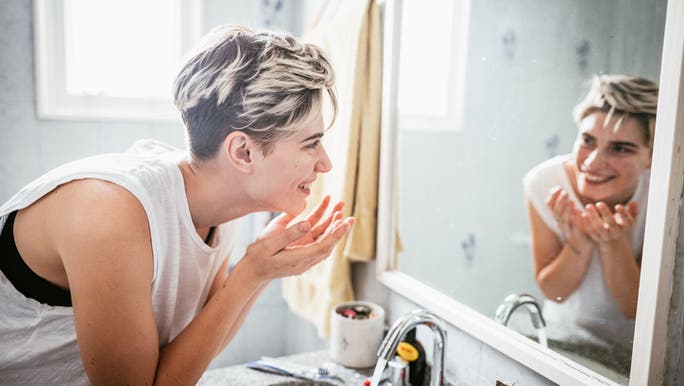 A woman with short blonde hair is washing her face and smiling at herself in the bathroom mirror