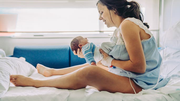 Holding her newborn baby while smiling on a hospital bed, a woman wonders how to stop hiccups in newborns. 