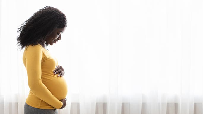 A woman wears a yellow sweater and cradles her baby bump in front of a white sheer curtain, thinking about her dry lips during pregnancy.
