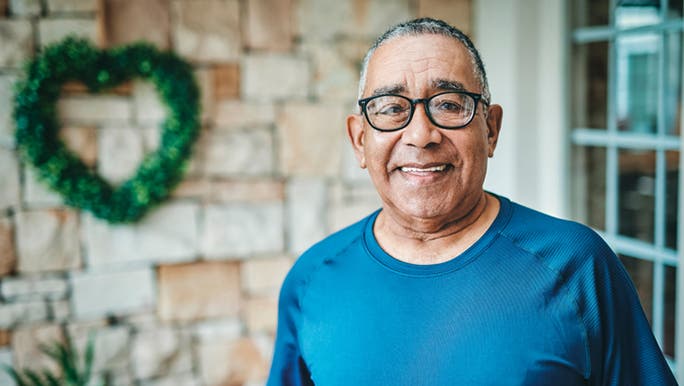 An older man wearing a blue T-shirt and glasses is standing outside a house and looking at the camera, smiling