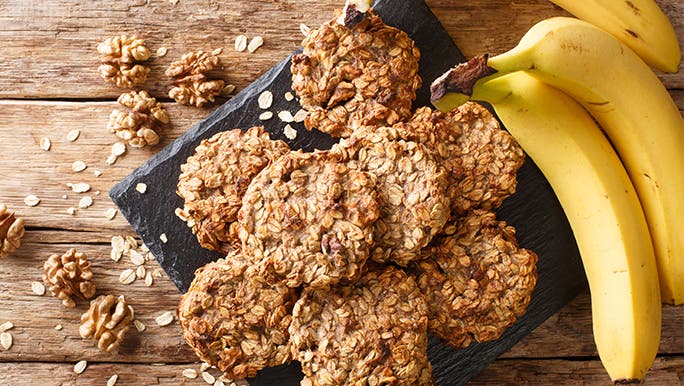 Top view of homemade banana-oat biscuits on a slate. The table is rustic and wooden and there are brainfoods like walnuts and oats scattered around. 