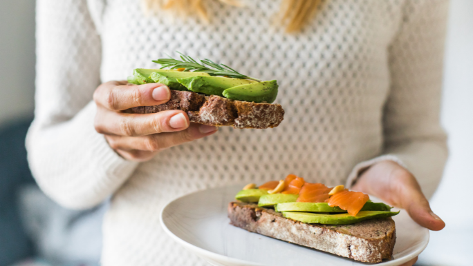 A woman is eating whole grain toast topped with avocado and salmon