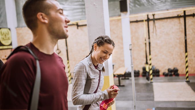 Two people smile as they walk through a gym with shoulder bags, discussing what to bring to the gym.