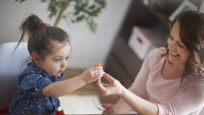 Kids can develop a healthy relationship with vegetables like this child looking curiously at a piece of vegetable that her mum is handing to her.