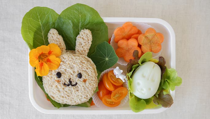 From above, an open lunch box containing a sandwich shaped like a rabbit and fresh, chopped veggies. It seems as though it may have been prepared for a fussy toddler. 