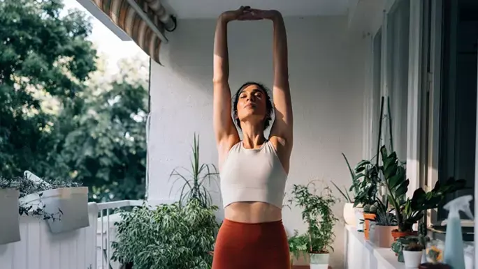 Woman in orange and white active wear standing on her balcony stretching her arms overhead