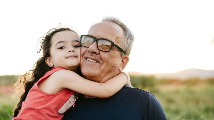 A little girl wraps her arms around her grandpa’s neck, there’s rolling green hills behind them and the light looks warm. 