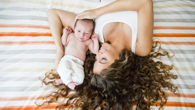 A lady is flopped on a bed with her curly hair around her face like a halo. She has a newborn who is cuddling into her armpit.