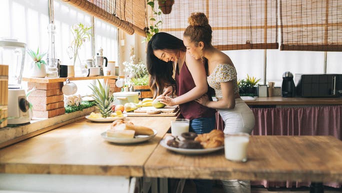 Two women are hugging and smiling as they prepare food in a kitchen with timber benchtops