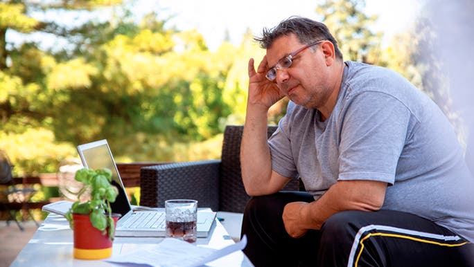 A large man wearing grey T-shirt and glasses is sitting in front of a laptop looking stressed