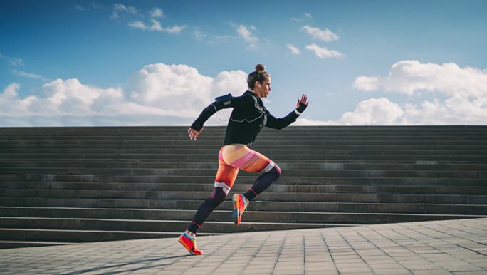 From the side, a woman wearing bright activewear sprints up a concrete slope. There are concrete stairs behind her and above them, a blue sky. 