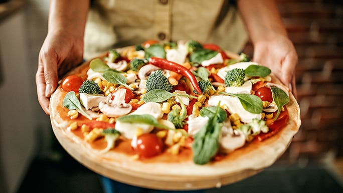 Two hands hold a pizza that’s covered in mushrooms, broccoli, tomato and spinach. 