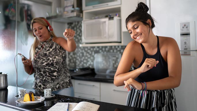A mother and daughter work on their fitness by dancing in their kitchen while preparing food.