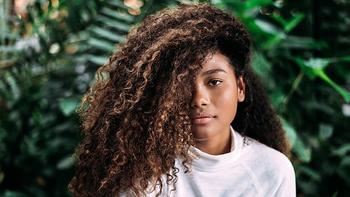 A woman with curly hair sits in front of some greenery. Her hair is covering half of her face. 