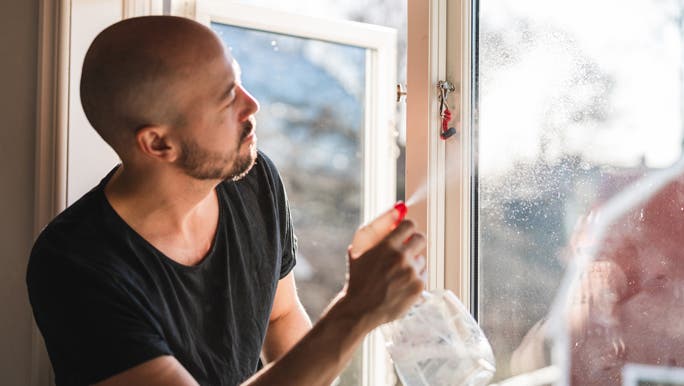 A man sitting on the window sill, cleaning the windows with spray cleaner and toilet paper on a sunny springtime day.