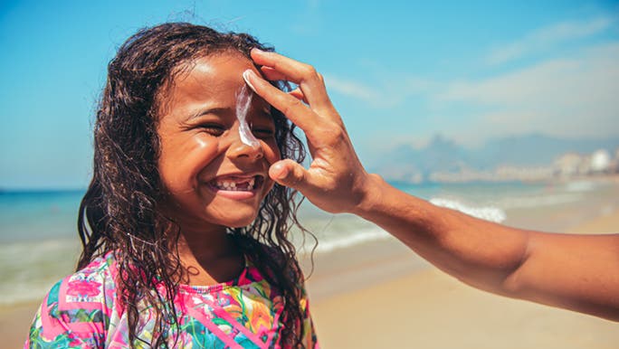 A little girl smiles at the beach as someone puts sunscreen on her face, hoping SPF 15 is enough.