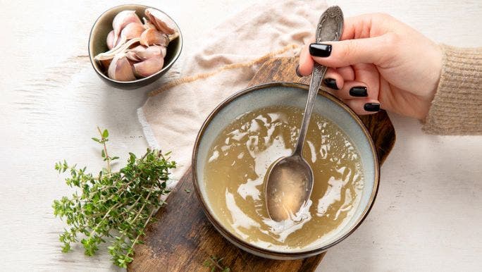 A woman’s hand is pictured holding a spoon in a bowl of bone broth