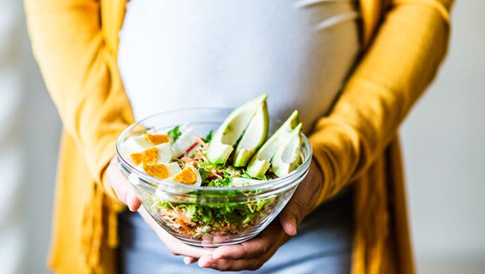 Pregnant woman wearing a yellow cardigan holding a salad bowl containing foods that may be considered good choices for someone following a diet to support fertility