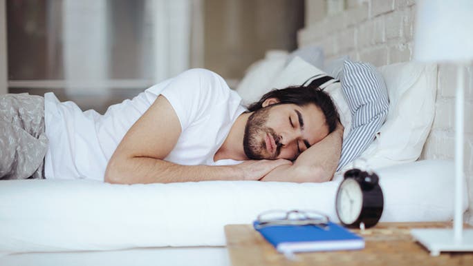 A dark-haired man wearing a white T-shirt is asleep in bed with an alarm clock on a bedside table beside him
