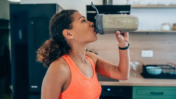 A woman in a bright orange racerback drinks the best pre workout powder from a protein shaker bottle.
