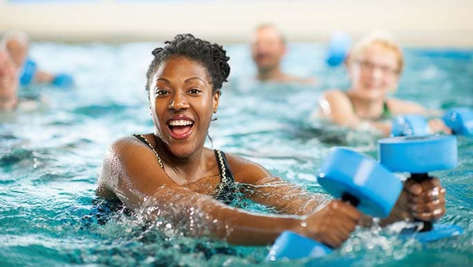 A woman in a pool holds weights in her hand while smiling as she thinks about whether swimming is good for pelvic floor muscles. 