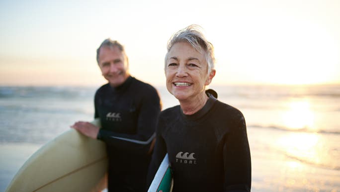 A healthy, ageing couple are standing on the beach at sunset. They are holding surf boards and wearing wetsuits.