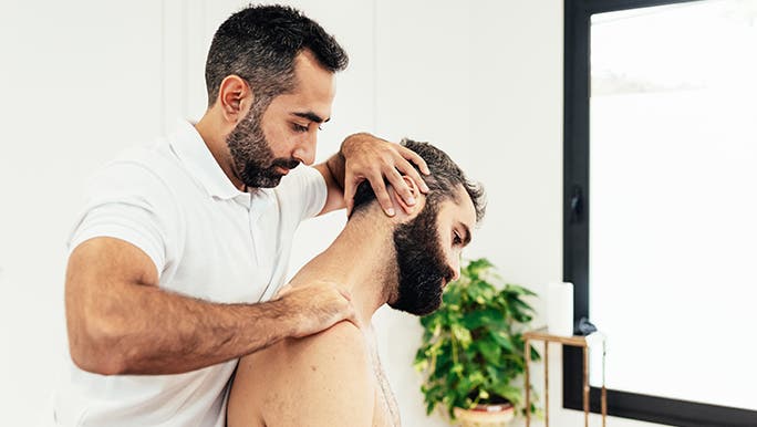 A remedial massage therapist is working on a man’s neck. They both have highly groomed beards. 