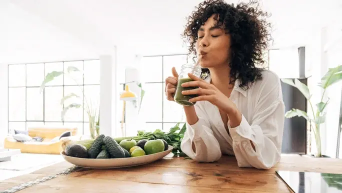 Young woman with curly black hair sipping a green juice in the kitchen
