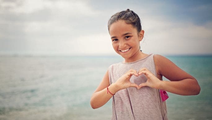 A young girl is standing on a beach and making a love heart shape with her hands in front of her chest