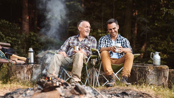 Two men sitting in camping chairs in front of a fire while camping in the outdoors