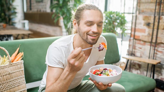 A young Caucasian man wearing a white T-shirt is sitting on a green sofa and smiling as he looks at a bowl of healthy food and practices mindful eating