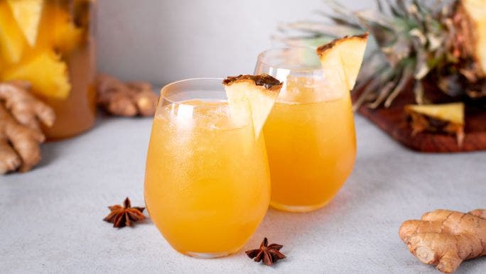 Two glasses of fresh pineapple, ginger and turmeric juice are sitting on a table with pineapple and ginger in the background and foreground