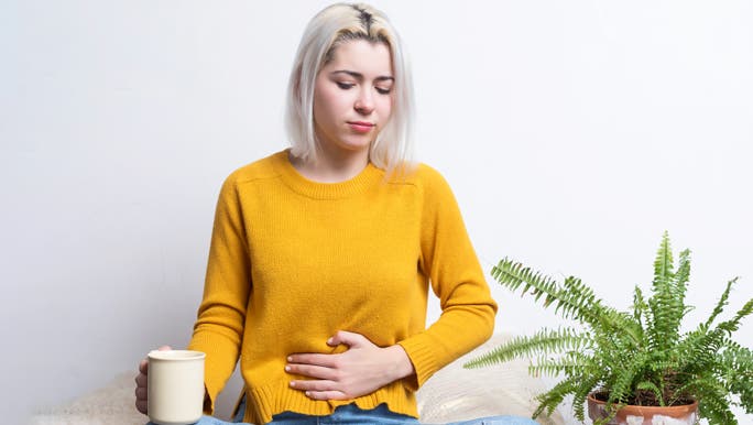 Young female sitting at home holding a cup of tea while putting her hand in her stomach