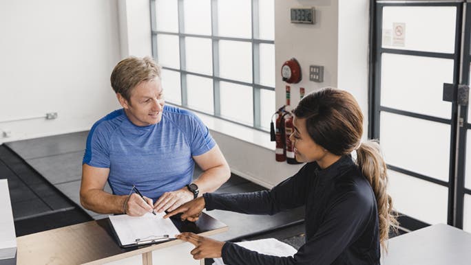 From above, a gym worker helps a man fill out paperwork for his gym membership.