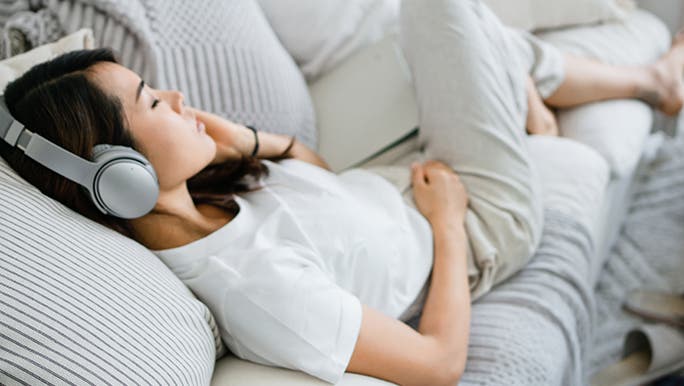 A lady takes a moment to relax on her couch with earphones on. It’s a bright room and her and the couch are both dressed in neutral colours. 