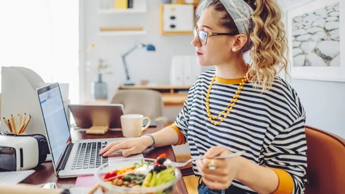 Woman in a striped t-shirt eating a salad and working from home on a laptop