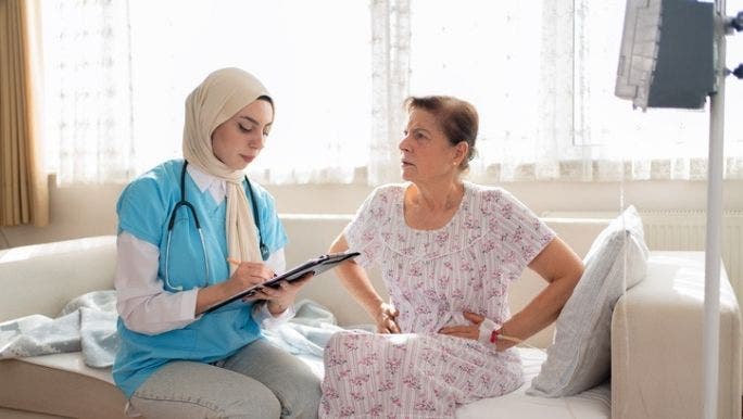 A medical professional wearing a headscarf is giving a senior woman a medical consultation as they sit together on a sofa