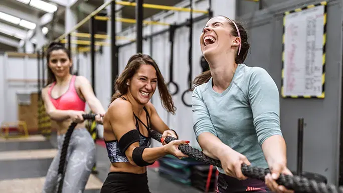 Three women are dressed in fitness gear at a gym. They are standing upright and pulling a gym battle rope while laughing and smiling.