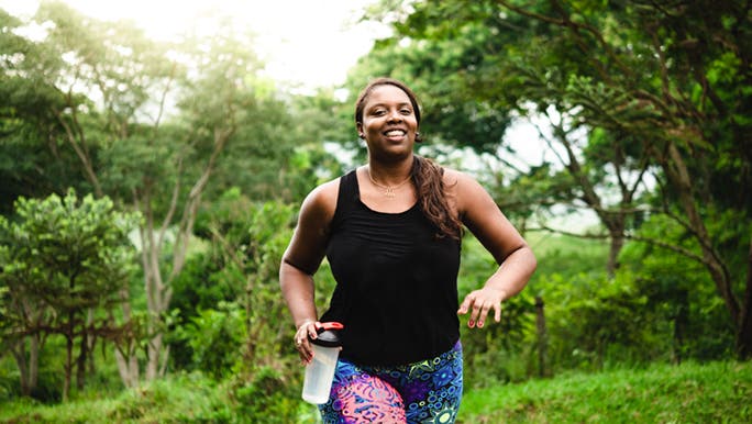 A woman smiles as she jogs up a hill, she is carrying a shaker full of water. It looks like one that comes with weight loss diet shakes. 