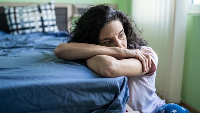 A dark-haired woman is sitting on the floor beside her bed with her head in her arms looking tired