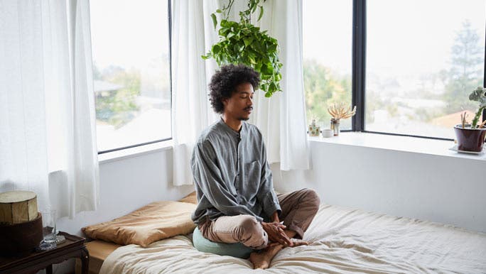 A man in a light filled bedroom sits on his bed meditating as he makes his mental health a new years resolution. 