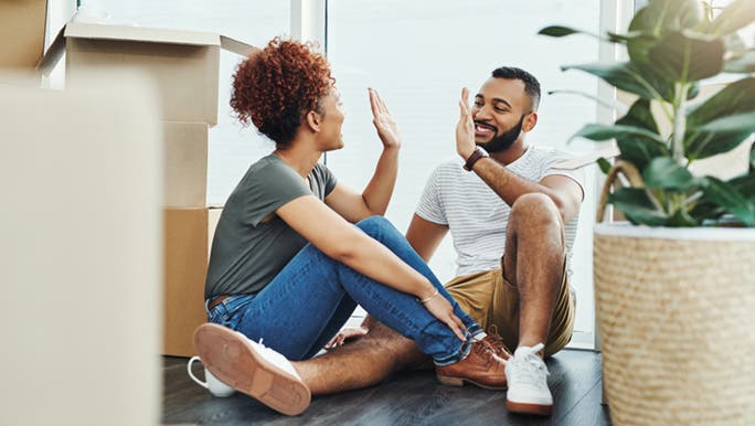 A couple sits on a floor surrounded by moving boxes, they are about to give each other a high five. It’s hard to tell by the picture what their love language is.