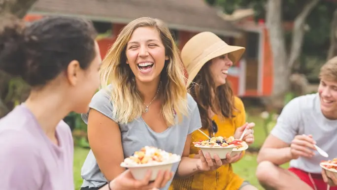 Four friends sit outside eating superfood smoothie bowls while laughing and talking