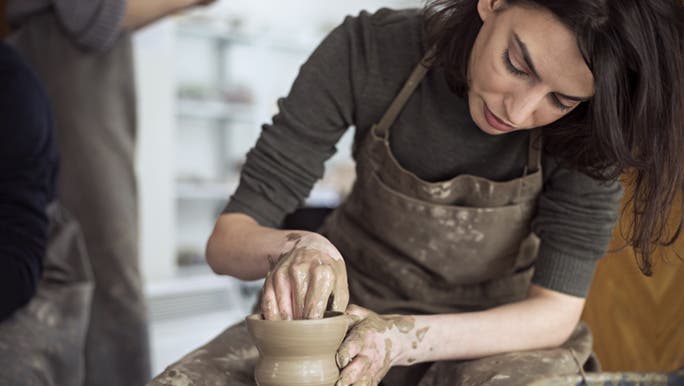 A woman is doing pottery on a wheel, she may be learning a new hobby to improve her mental wellbeing. 