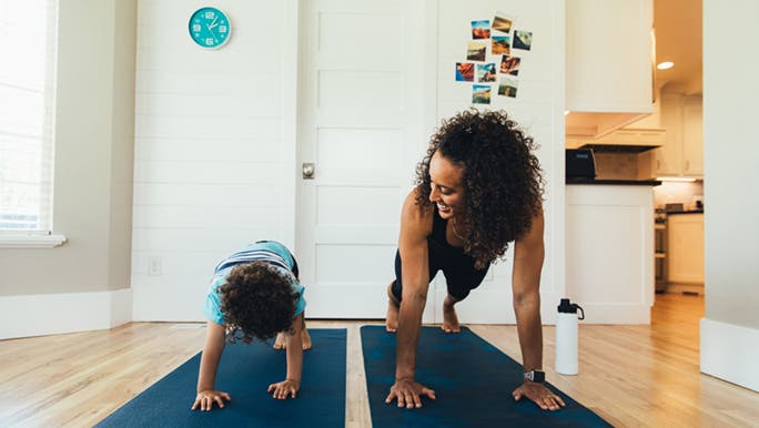 A woman and her child are exercising for mental health. They have two navy yoga mats rolled out on the floor of their home and are doing the plank position. 