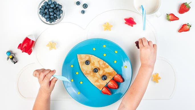 From above, a child’s breakfast on a blue plate. It’s a pancake shaped like a rocket ship with strawberries and blueberries on it. There is also a cup of milk with a straw and two arms of a child. 